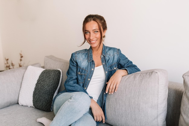 Fulllenght photo of smiling lovely woman with dark hair and wonderful smile wearing denim shirt sitting on sofa and posing at camera in modern stylish apartment