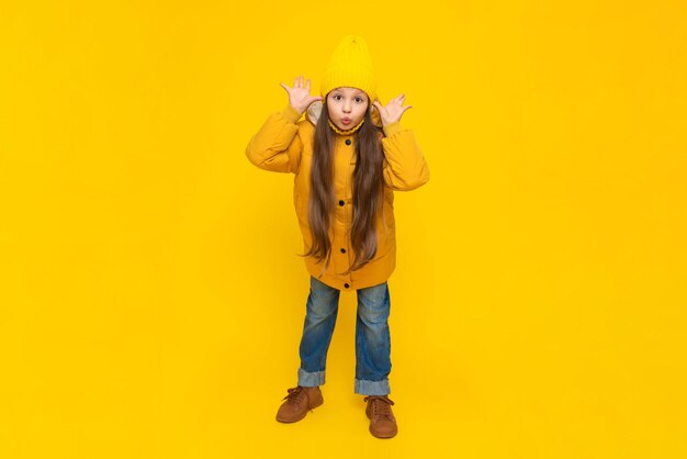 A fulllength little girl in a down jacket and a hat puffed out her cheeks The child is happy about the onset of autumn Warm clothes for children Yellow isolated background