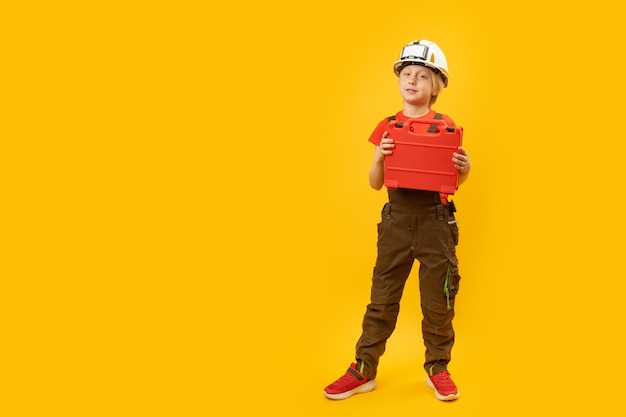 Fulllength portrait of boy imitating worker Child wears helmet with holds toolbox isolation on yellow background Copy space mock up