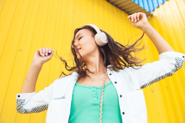 A fulllength view of a beautiful cheerful girl jumping while listening to a melody isolated against a bright yellow background a young woman with headphones dancing on the street generation z