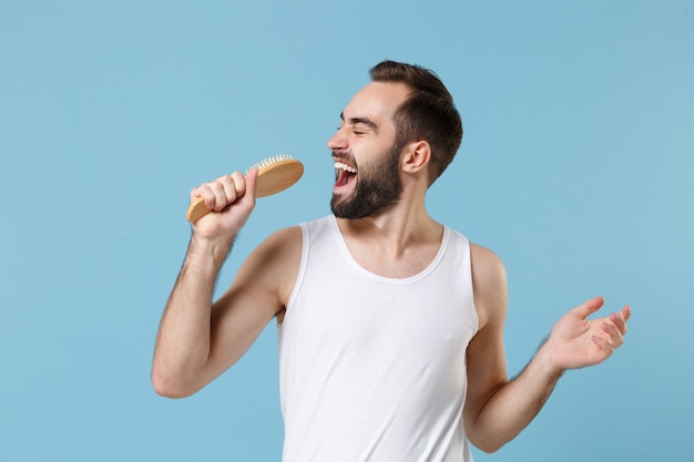 Fun bearded man 20s years old in white shirt sing hold comb like microphone isolated on blue pastel wall,  portrait. Skin care healthcare cosmetic procedures concept. 