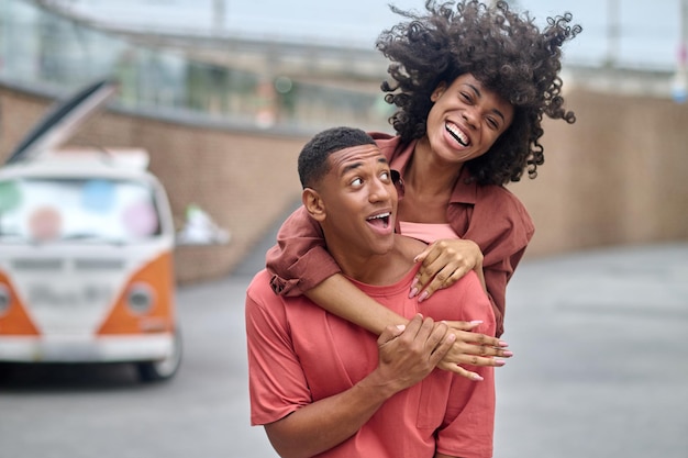 Fun case. Young energetic african american woman with wind in curly hair on back of surprised smiling man with big eyes having fun outdoors during day