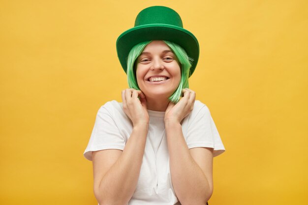 Fun festivities on St Patricks Day Cute charming woman with green hair wearing leprechaun hat standing isolated over yellow background looking at camera with toothy smile