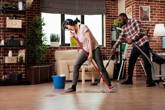 Fun housewife cleaning with husband, listening to music with headphones singing and dancing happily. Modern married couple enjoying together taking care of household chores.