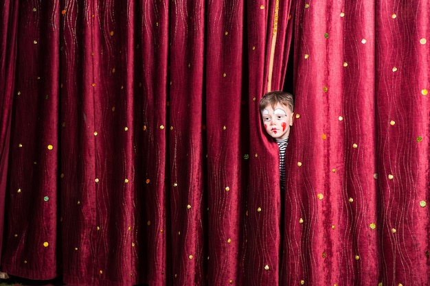 Fun little boy in makeup waiting for his acting cue poking his head out between the curtains as he waits to make his entrance on stage during the performance