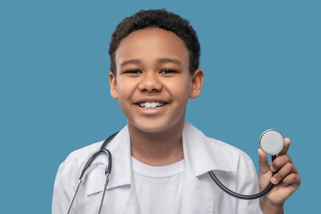 Fun mood. Laughing african american boy of primary school age in medical gown with stethoscope on background in studio