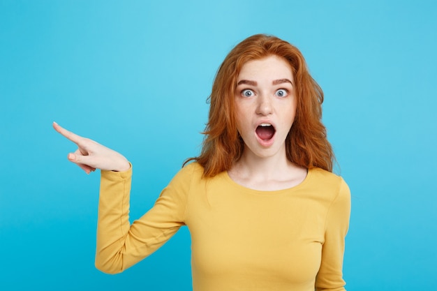 Fun and people concept  headshot portrait of happy ginger red hair girl with pointing finger away and shocking expression pastel blue wall copy space