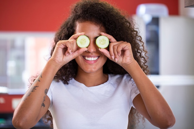 Fun portrait of beautiful black african model holding a cucumber slice to her eye, rejuvenating skin care regime treatment facial beauty concept.