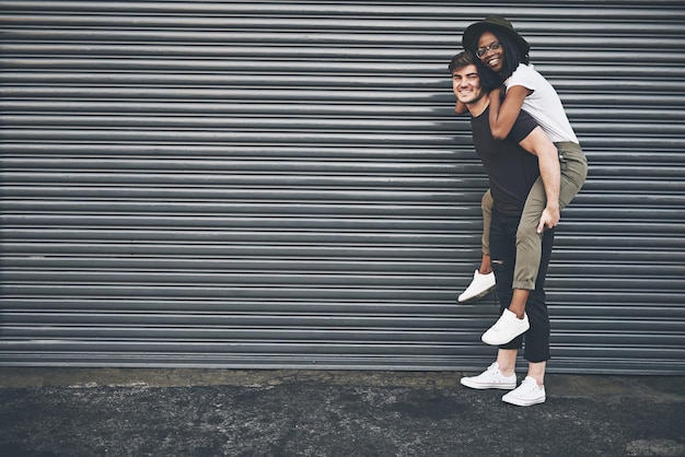 Fun trendy and happy interracial couple enjoying a piggyback ride together outside against an urban background Lovers bonding and being playful Smiling and relaxing while walking outdoors