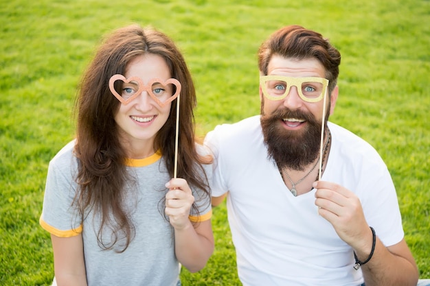 Fun Valentines props Bearded man and sexy girl smiling with fancy party props on green grass Funny couple holding heart shaped photobooth props on stick Happy family celebrating with glasses props