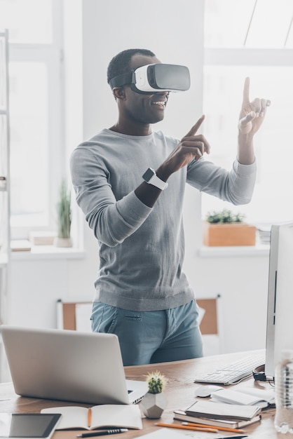 Fun with virtual reality headset. Handsome young African man in VR headset gesturing and smiling while standing in creative office