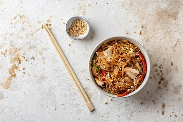 Funchose salad with chicken and vegetables in bowl with wooden chopsticks on textured background top view with text space Asian food menu