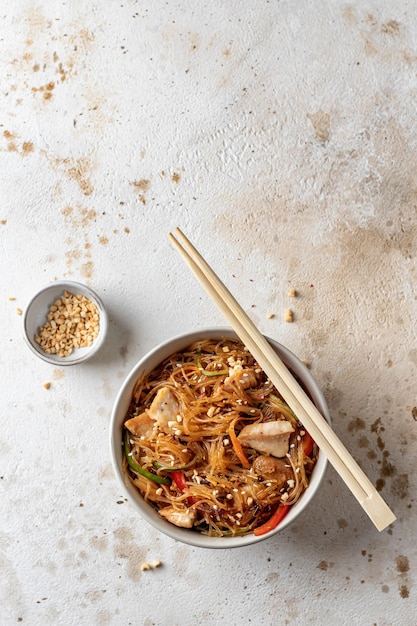 Funchose salad with chicken and vegetables in bowl with wooden chopsticks on textured brown background with copy space Food menu Top view