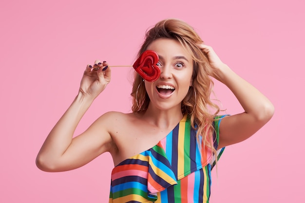 Funky positive cheerful attractive lady with her stylish trendy wave curly hair, holds a sugar sucker on stick isolated on pink