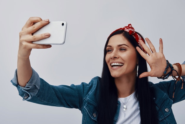 Funky young woman in bandana making selfie and smiling while standing against grey wall