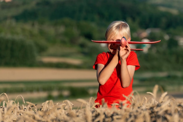 Funny 7 year boy with blond hair play with red plane and standing in ears of ripe wheat Air travel with children