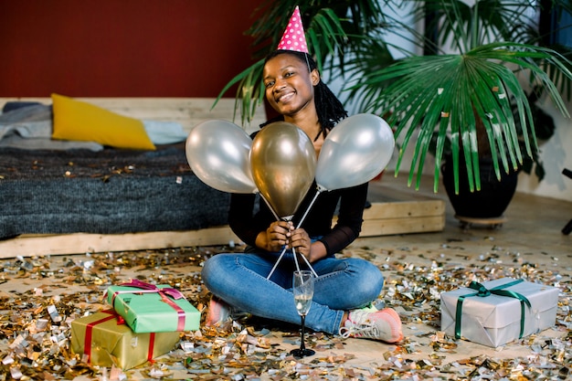 Funny African American girl in birthday hat and with air balloons sitting on the floor with present boxes gifts and confetti. Celebration, party concept