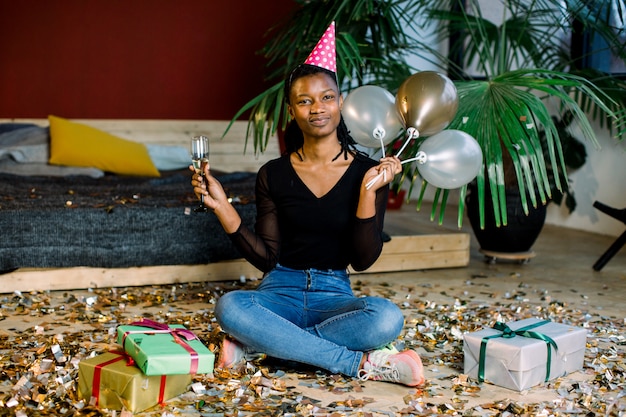 Funny African American girl in birthday hat and with air balloons sitting on the floor with present boxes gifts and confetti. Celebration, party concept