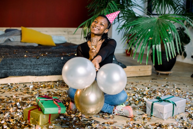 Funny African American girl in birthday hat and with air balloons sitting on the floor with present boxes gifts and confetti. Celebration, party concept