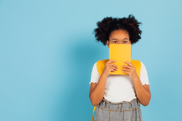 Funny african american schoolgirl covering her face with yellow book on blue background back to school concept