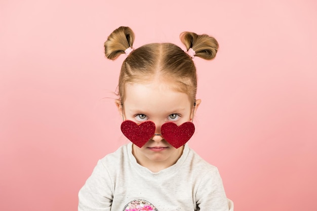 Funny angry little blonde girl in red heart shape sunglasses on pink background. Valentines Day or Mothers Day concept.