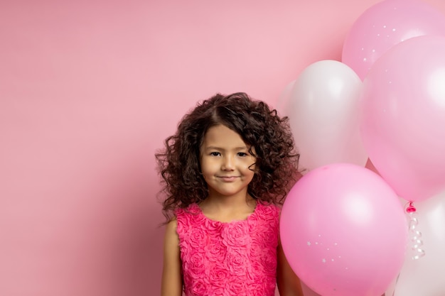 Funny attractive kid with curly hair, standing near pink and white air balloons on birthday party, wearing lovely dress, smiling , isolated on rosy wall with copy space. Happy childhood, holiday.