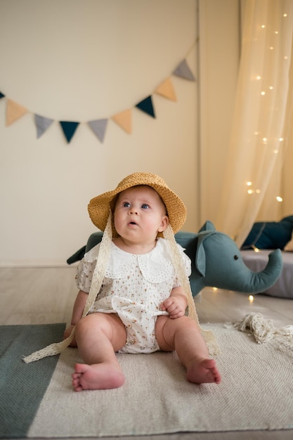 Funny baby girl in a bodysuit and a straw hat is sitting on the floor in the nursery