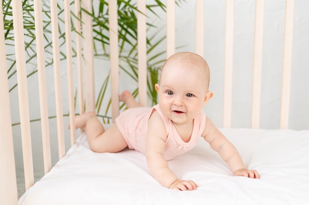 Funny baby girl in a crib in a pink bodysuit for six months on a white cotton bed lies on her stomach and smiles, woke up in the morning or goes to bed