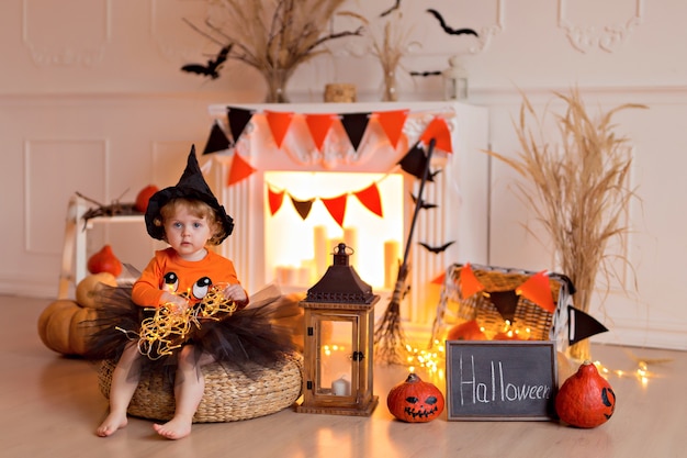 Funny baby girl in halloween witch costume with pumpkin jack and broom indoors