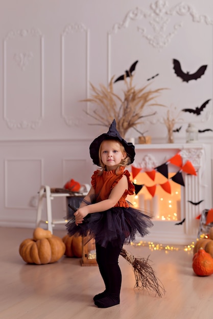 Funny baby girl in halloween witch costume with pumpkin jack and broom indoors