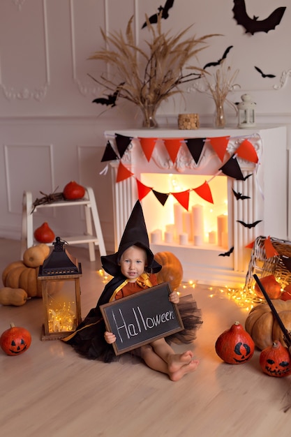 Funny baby girl in halloween witch costume with pumpkin jack and broom indoors