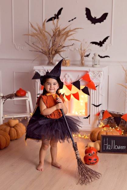 Funny baby girl in halloween witch costume with pumpkin jack and broom indoors