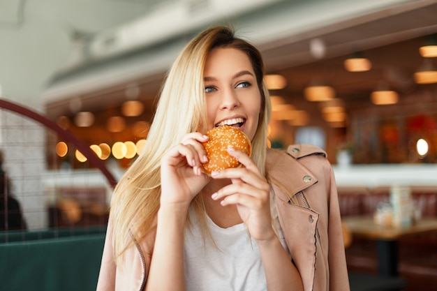 Funny beautiful young woman in a pink jacket eating a hamburger in a cafe