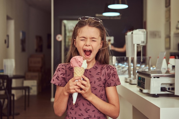 A funny beauty little girl in a fashionable dress eating strawberry, standing in an ice cream parlor.