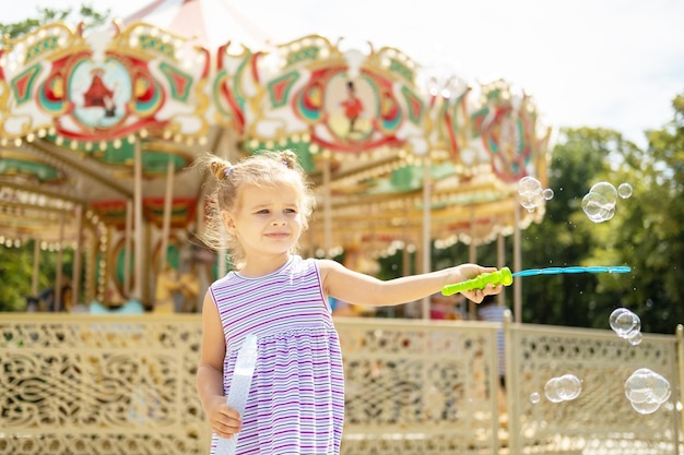 Funny blonde girl in colorful dress playing with soap bubbles at the carnival