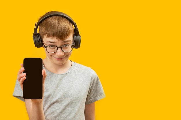 Funny boy in glasses and large black headphones shows phone with empty black screen on yellow background Copy space Mock up