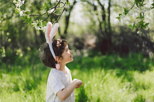 Funny boy with eggs basket and bunny ears on easter egg hunt in sunny spring garden