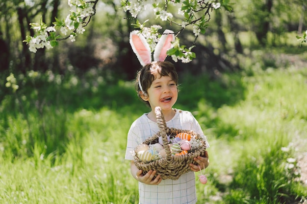 Funny boy with eggs basket and bunny ears on Easter egg hunt in sunny spring garden