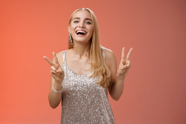 Funny carefree charming caucasian blond female model in silver stylish glamour dress show peace victory gesture smiling broadly have fun celebrating birthday party, standing red background.