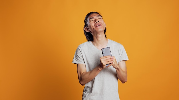 Funny carefree man singing songs on camera, using smartphone as microphone and fooling around. Confident young guy acting silly or funky, enjoying music and audio in studio.