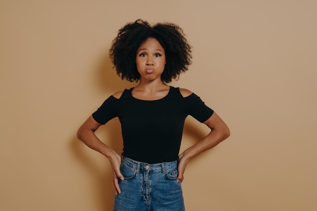 Funny carefree young dark skinned female puffing out her cheeks against nude studio wall background