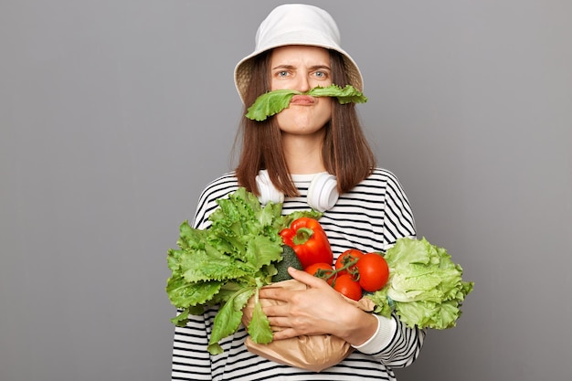 Funny Caucasian woman holding organic vegetables standing isolated over gray background making mustache with green lettuce looking at camera