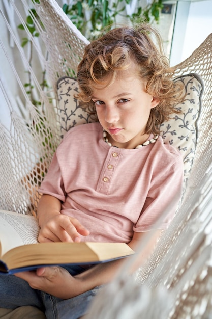 Funny child in casual clothes resting in knitted white hammock while reading novel and looking at camera on balcony at home on sunny weekend day