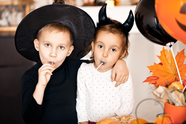 Funny child girl and boy in witch and evil costumes for Halloween eating candies lolly pops and have fun.