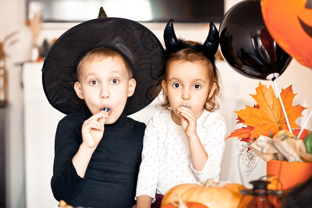 Funny child girl and teen boy in witch and evil costumes for Halloween party eating candies lolly pops and have fun