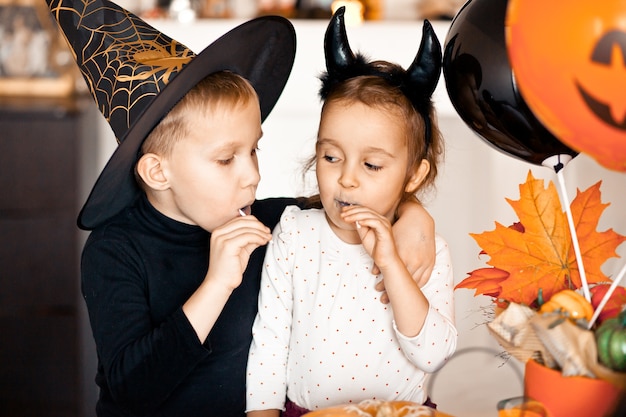 Funny child girl and teen boy in witch and evil costumes for Halloween party eating candies lolly pops and have fun