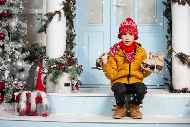 Funny child holds old skates and sits on the porch of a house decorated with Christmas tree