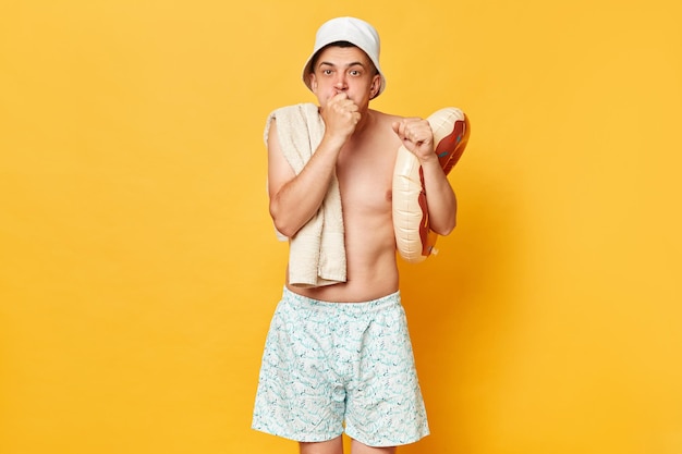 Funny confused man man wearing shorts swimsuit and panama holding donut rubber ring isolated on yellow background clenched fist looking at camera