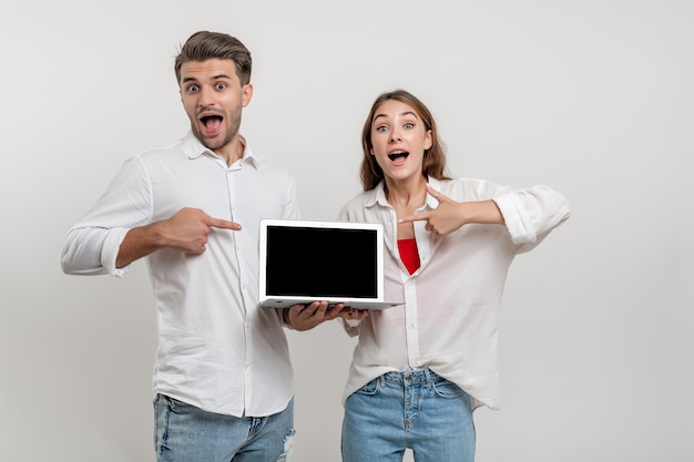 Funny couple friends pointing index fingers on laptop computer with empty screen isolated over white