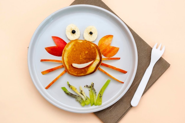 Funny crab face shape snack from pancakeapplesbananakiwi on plate Cute kids childrens baby's sweet dessert healthy breakfastlunch food art on beige backgroundtop view flat lay